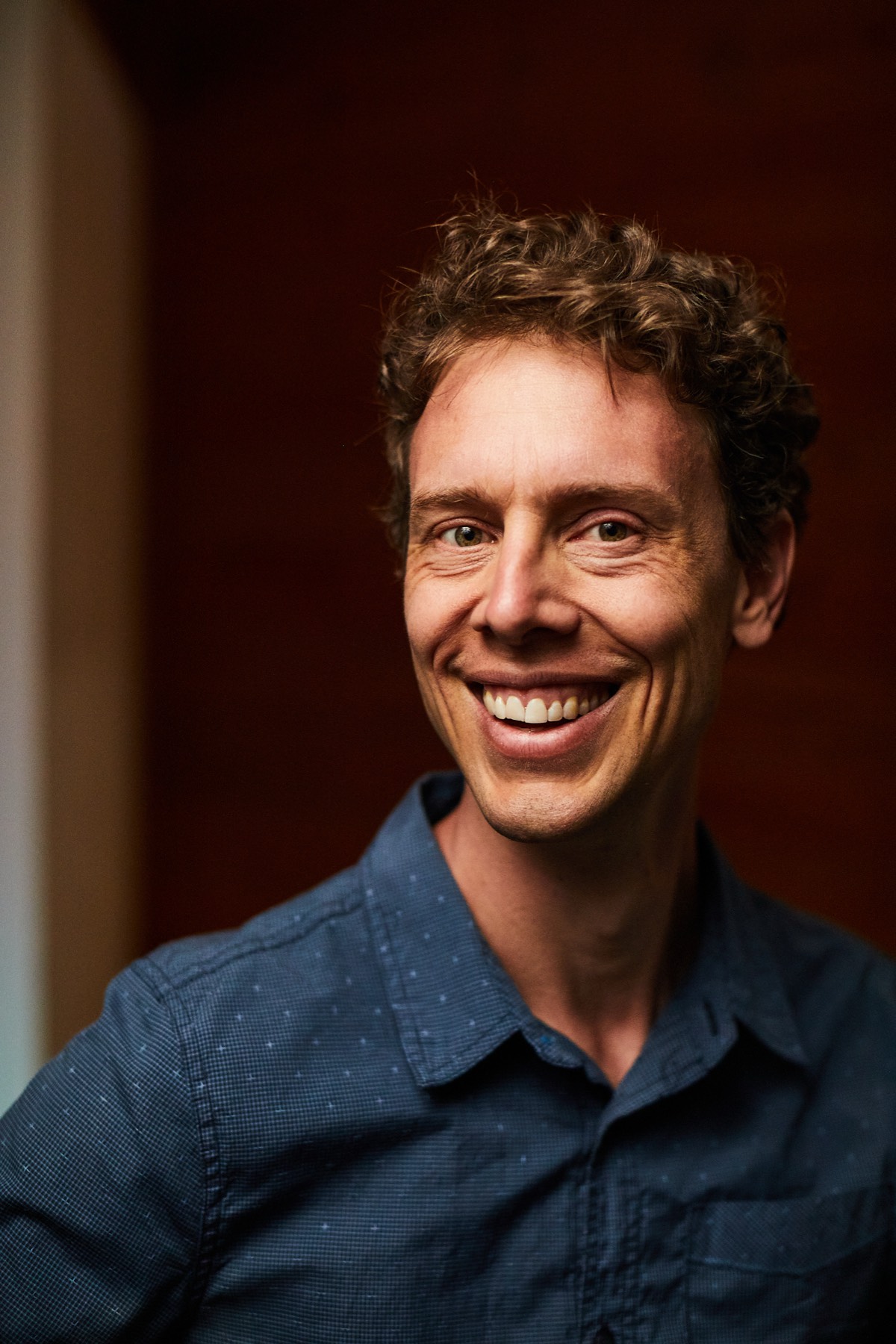 Mark Andreas with a warm, direct smile to camera against a deep red-brown background.