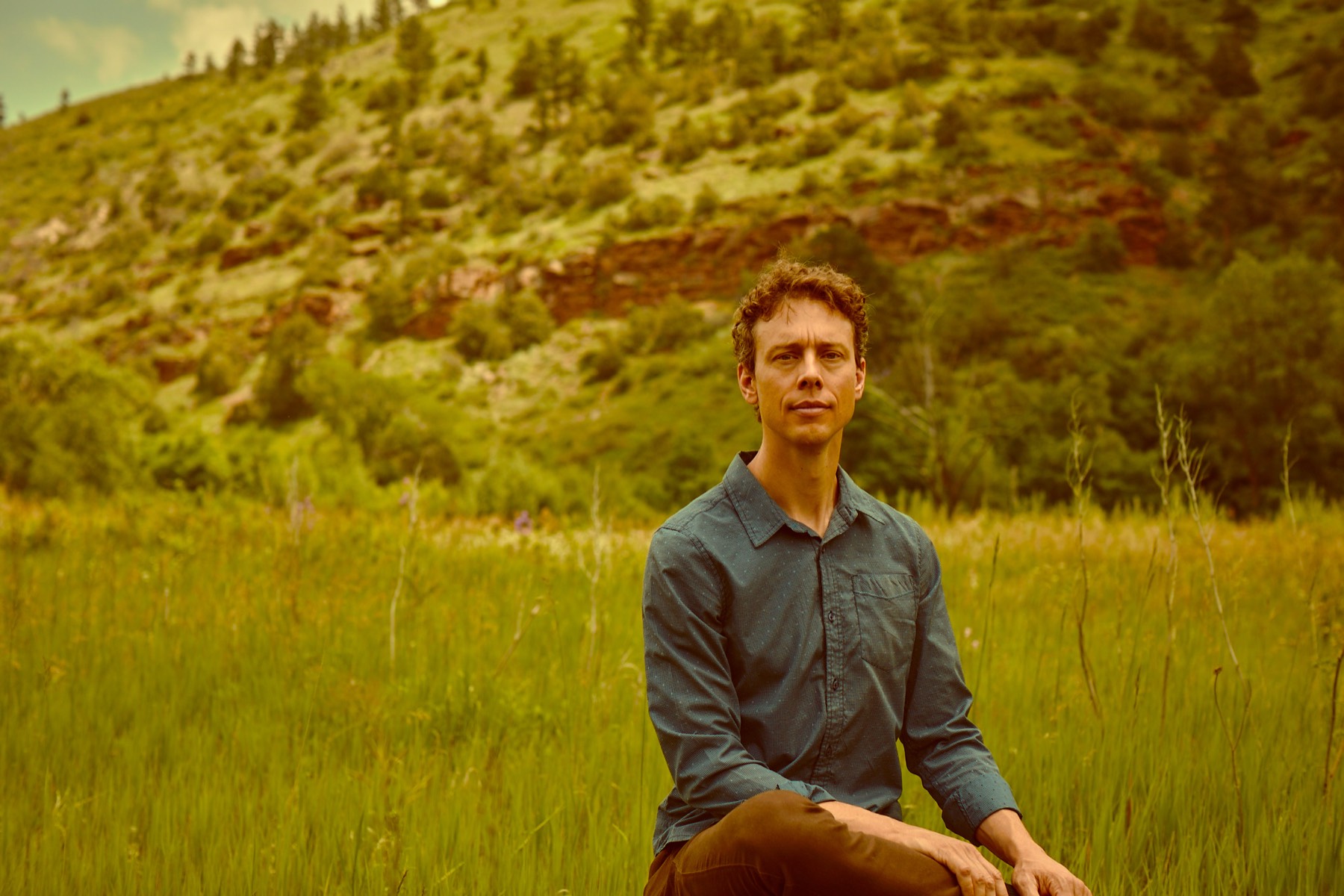 Mark Andreas seated in a meadow, a red-rock hillside behind him, calm and contemplative.